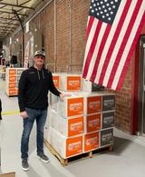 Man stands by stacked "Hunger Fight" boxes on a pallet, next to a large American flag indoors.