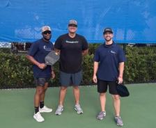 Three Alpha Foundations reps stand on an outdoor pickleball court, holding paddles and smiling at the camera.