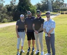 Four men in golf attire stand together on a sunny golf course, smiling at the camera.