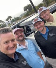 Four smiling men in golf attire pose together outdoors near golf carts on a cloudy day.