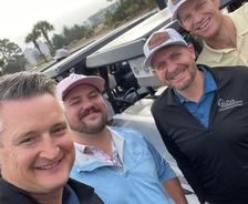 Four smiling men in golf attire pose together outdoors near golf carts on a cloudy day.