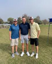 Three men in golf attire smiling and standing on a grassy golf course under a cloudy sky.