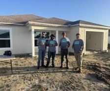 Four men in matching shirts stand in front of a new house under construction on a sunny day.