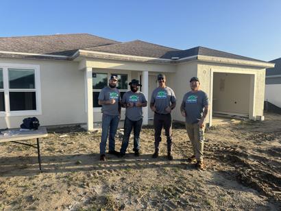 Four men in matching shirts stand in front of a new house under construction on a sunny day.