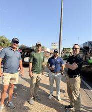 Four men in casual clothes stand outside on a sunny day, smiling, with cars and buildings in the background.