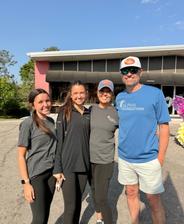 Four people smiling outdoors in casual clothes, standing together in front of a building on a sunny day.