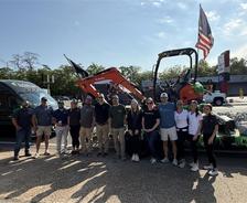 A group of people stands in front of a truck and equipment, with an American flag raised beside them.