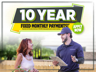 Man in uniform holding clipboard talks to smiling woman under a sign reading "10 Year Fixed Monthly Payments! Apply Now.