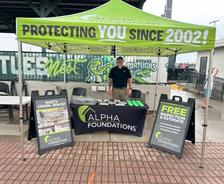 A man stands at an Alpha Foundations booth under a green tent with signs offering free foundation inspections at Daytona Tortugas Game Day 2026