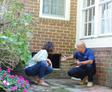 Two people discuss home maintenance, crouching by a brick wall with a small opening, surrounded by plants.