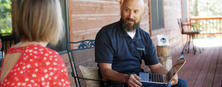 A man with a beard sits on a porch with a laptop, conversing with a woman in a red shirt.