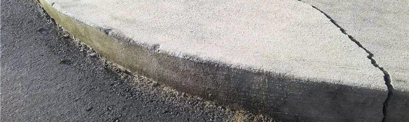 Cracked concrete sidewalk curb next to asphalt road and grass.