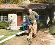 A man digs a trench in a garden near a house; pipes are placed beside the trench.