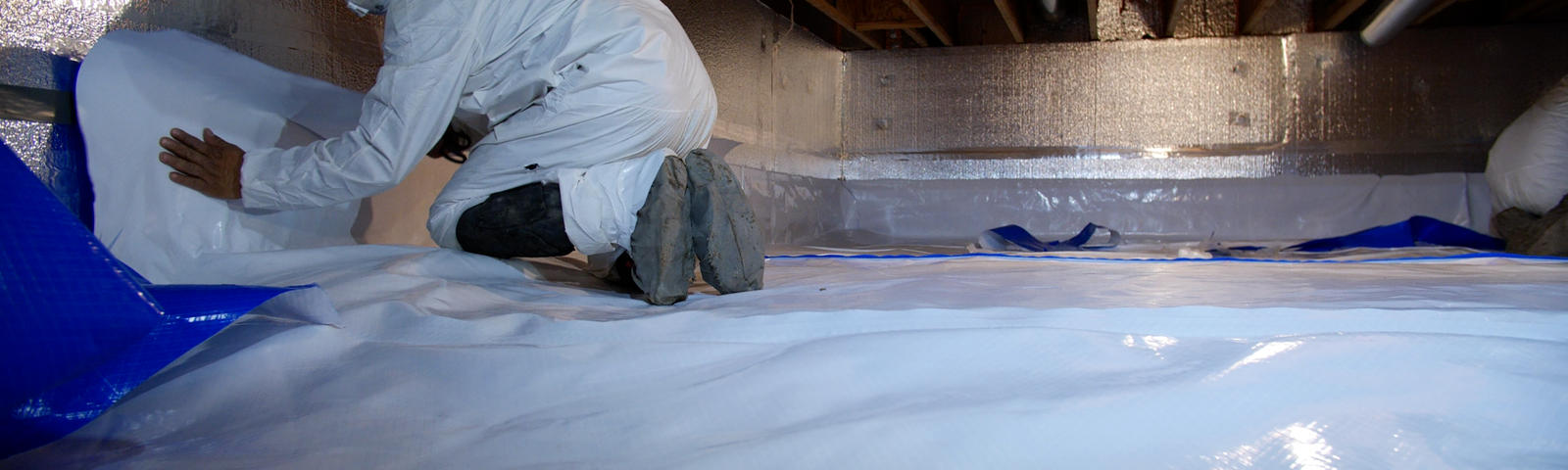 A person in protective gear installing a vapor barrier in a crawl space with wooden beams above.