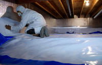 A person in protective gear installing a vapor barrier in a crawl space with wooden beams above.