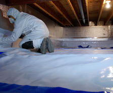 A person in protective gear installing a vapor barrier in a crawl space with wooden beams above.
