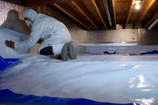 A person in protective gear installing a vapor barrier in a crawl space with wooden beams above.