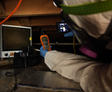 Person in protective gear inspecting equipment in a dark attic space with a flashlight and monitoring device.