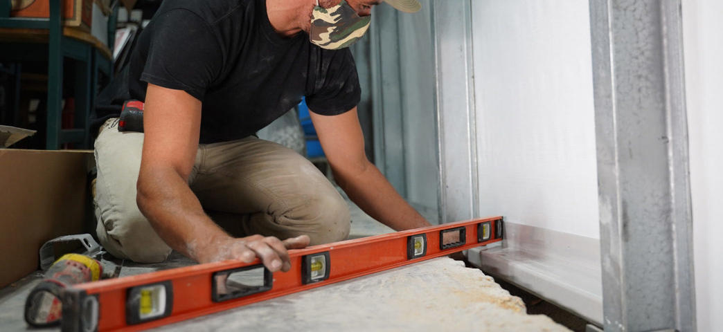 Person wearing a camo mask uses an orange level tool on a construction site floor.
