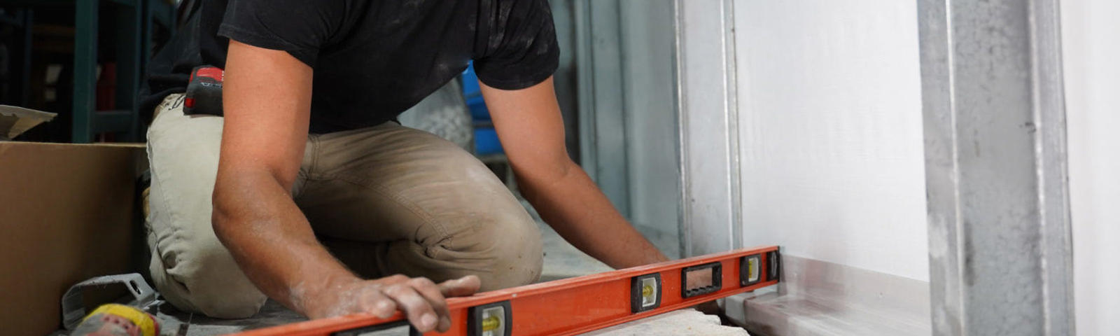 Person wearing a camo mask uses an orange level tool on a construction site floor.