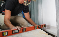 Person wearing a camo mask uses an orange level tool on a construction site floor.