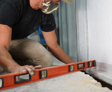 Person wearing a camo mask uses an orange level tool on a construction site floor.