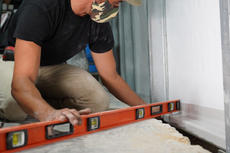 Person wearing a camo mask uses an orange level tool on a construction site floor.