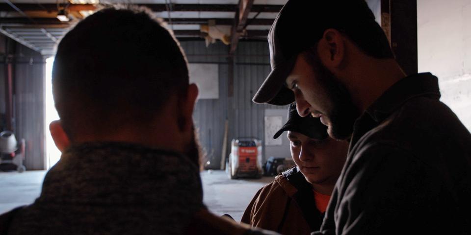 Three men in a dimly lit garage, two wearing caps, focused on something in front of them.