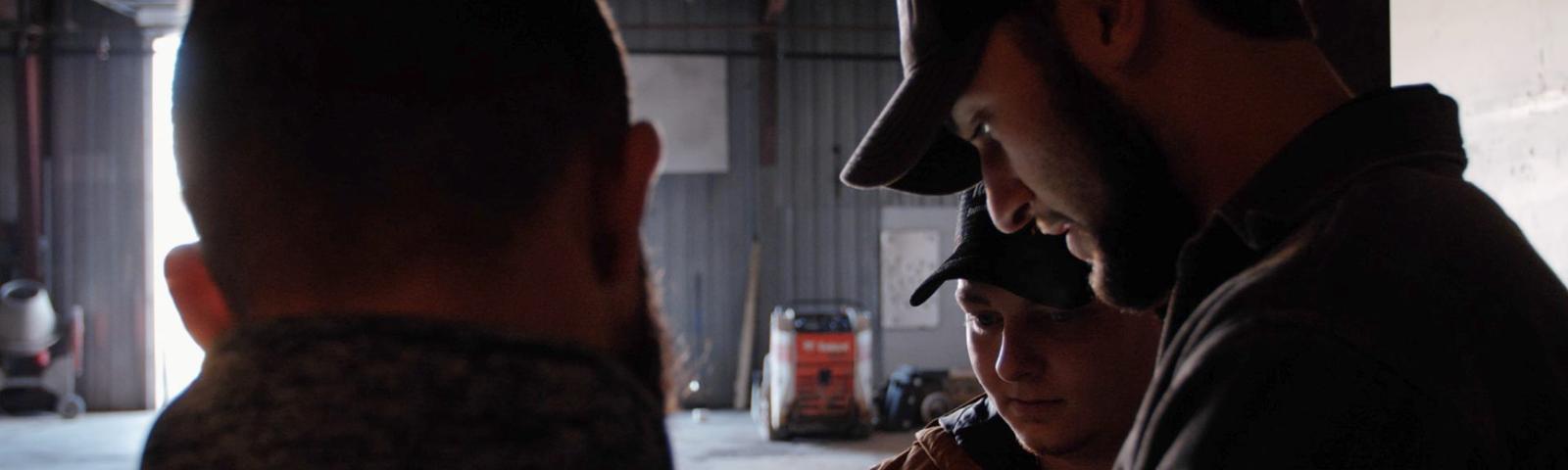 Three men in a dimly lit garage, two wearing caps, focused on something in front of them.