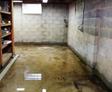 A basement with a concrete floor, visible water pooling, and walls lined with shelves holding various items.