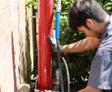 A worker operates a red hydraulic machine next to a brick wall and green bushes.