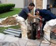 Two workers using equipment to install or repair a foundation outside a house, with pipes and plastic sheeting nearby.