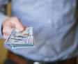 A person in a blue shirt holding a stack of US dollar bills extended toward the camera.