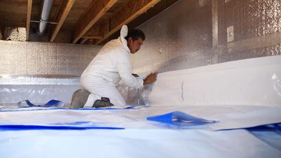 Worker in white overalls installing insulation in a crawl space or attic area.