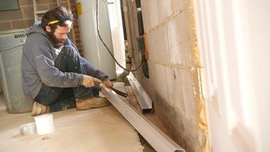 A man installing a metal drainage system along a brick wall in a basement.