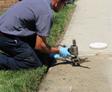 A person in uniform and gloves kneels on grass, working on a utility hookup near a sidewalk.