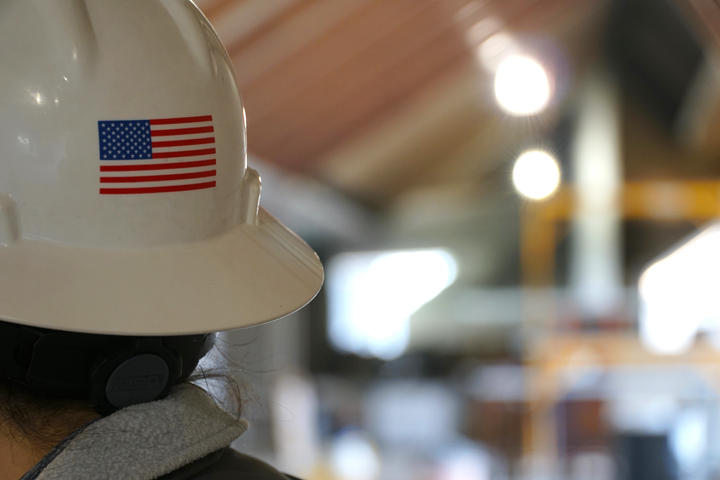 A person wearing a hard hat with a U.S. flag sticker, standing in an industrial setting with blurred background.