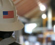 A person wearing a hard hat with a U.S. flag sticker, standing in an industrial setting with blurred background.