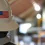 A person wearing a hard hat with a U.S. flag sticker, standing in an industrial setting with blurred background.