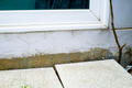 Close-up of a house wall with a window, exposed foundation, and concrete patio slabs.