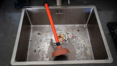 The plunger is sitting in the food-clogged kitchen sink.