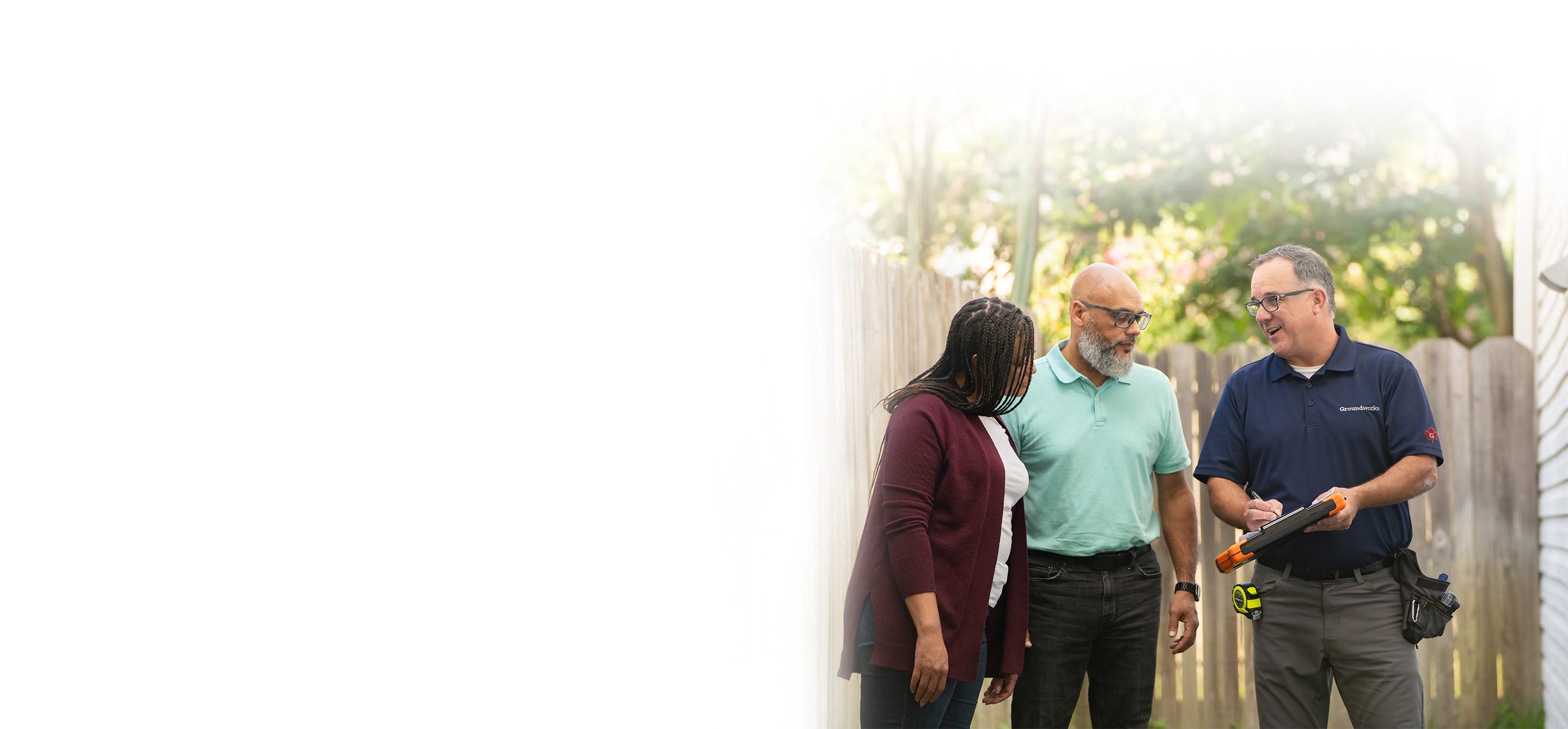 A group of three people standing outdoors, one holding a tablet while the others watch, with a wooden fence in the background.