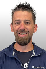 Smiling man with short hair and beard wearing a blue Groundworks jacket against a gray background.