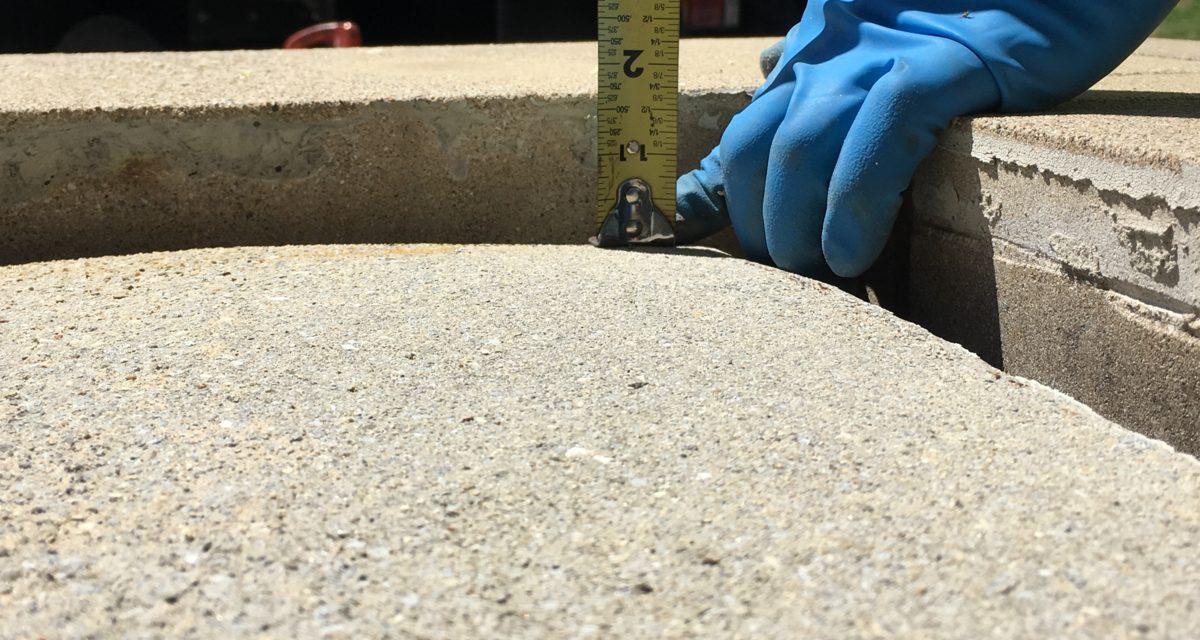 A gloved hand measuring the depth of an opening in a concrete surface with a yellow measuring tape.