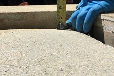 A gloved hand measuring the depth of an opening in a concrete surface with a yellow measuring tape.