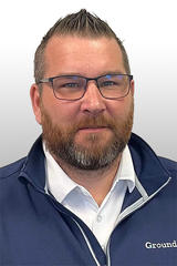 Man with glasses and beard wearing a navy jacket and white shirt, against a plain white background.