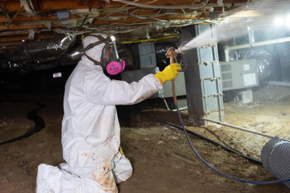 Person in protective gear spray-treating a crawl space under a house.
