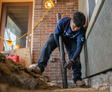 A worker installs a pipe at a construction site with exposed ground, wearing gloves and dark clothing.