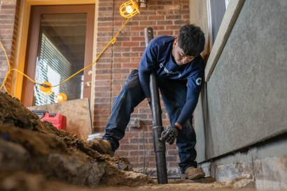 A worker installs a pipe at a construction site with exposed ground, wearing gloves and dark clothing.