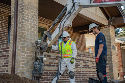 Two construction workers operate heavy machinery near a brick house, wearing hard hats and safety gear.